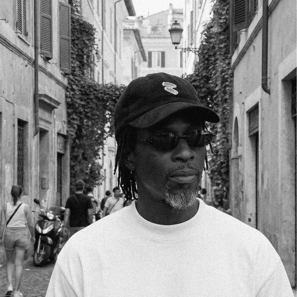 Man wearing a cap with two yellow bullet casings and sunglasses on a street with buildings and people in the background