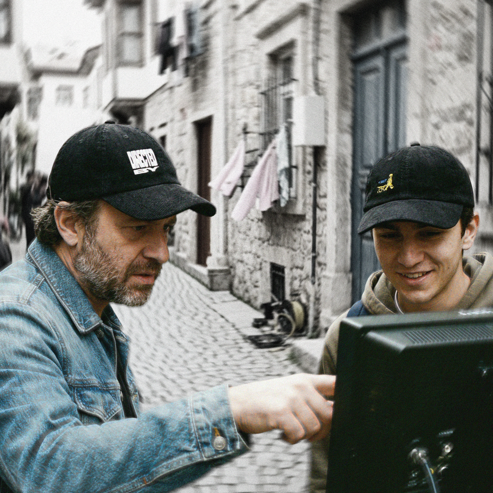 Two men in an urban setting, one wearing a black cap with a logo that reads: "DIRECTED BY" and the other wearing a black cap with a logo of a child and a dog looking at a screen.