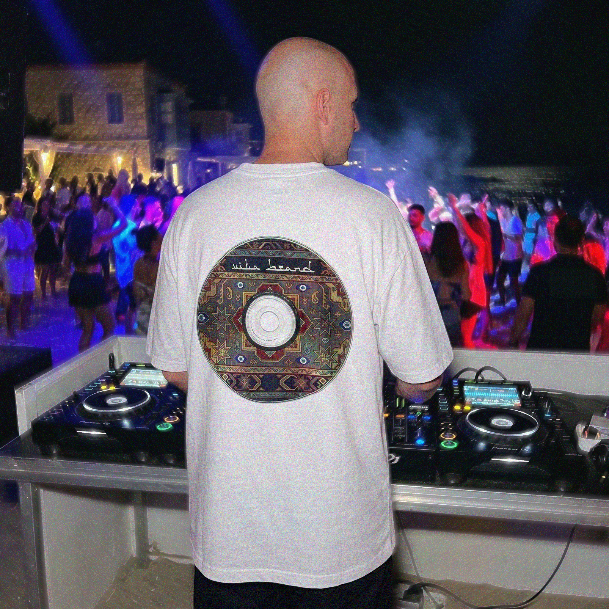 DJ at a music event with crowd and lights in the background wearing a white t-shirt with a turkish carpet CD logo that reads: "Vita Brand"