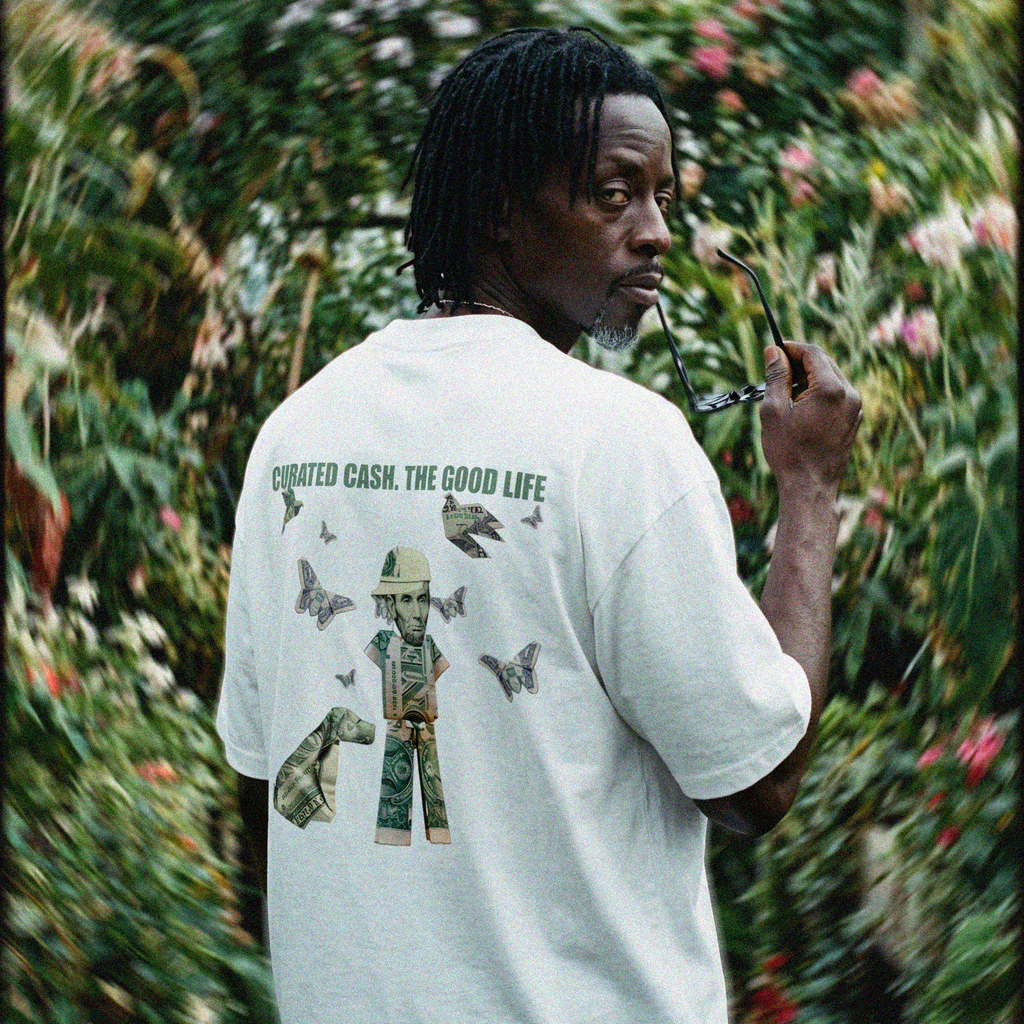 Man wearing a white t-shirt with a graphic design in a garden setting "CURATED CASH. THE GOOD LIFE."