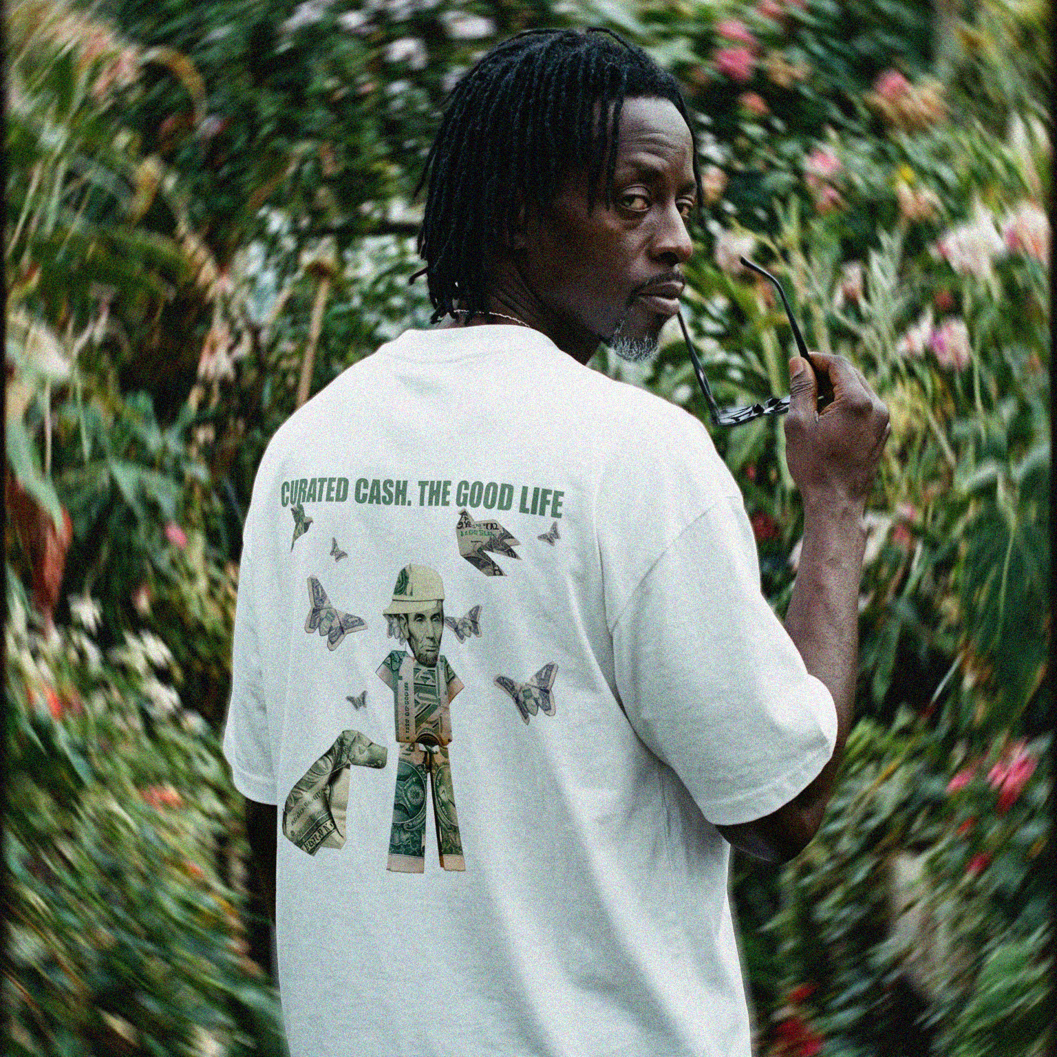 Man wearing a white t-shirt with a graphic design in a garden setting "CURATED CASH. THE GOOD LIFE."