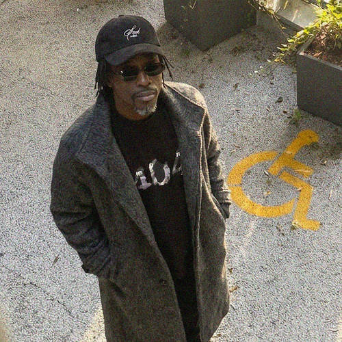 Tall black man wearing a black cap and sunglasses, standing on a concrete surface with graffiti. "404"