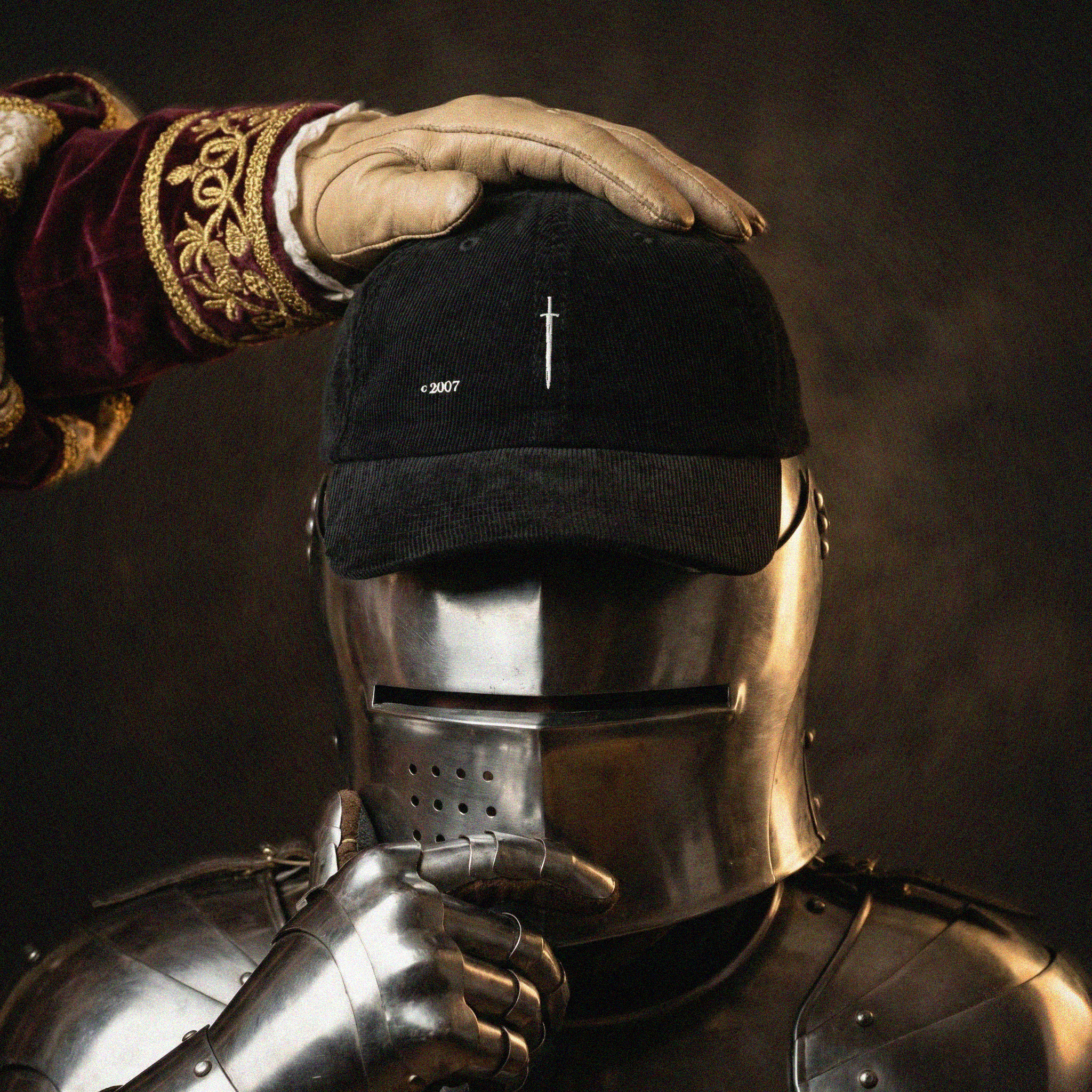 A black cap with a white sword logo being put on a knight by a king against a dark background